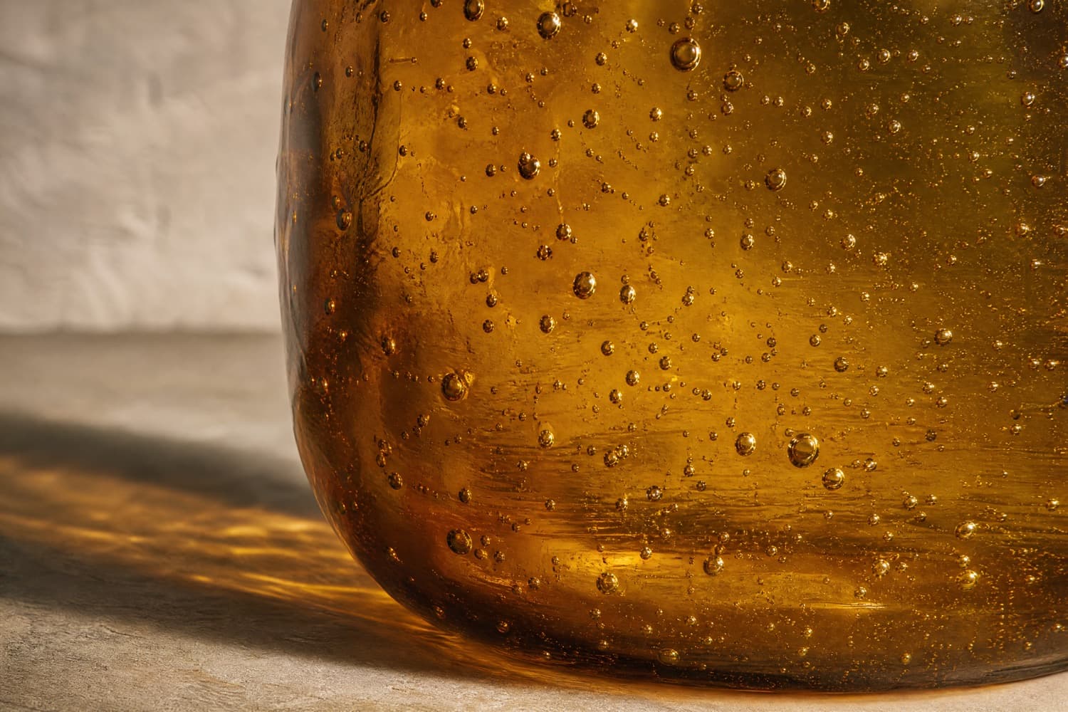 Extreme close-up of the wall of a mouth-blown amber glass, backlit so a scatter of trapped air bubbles reads as bright points.