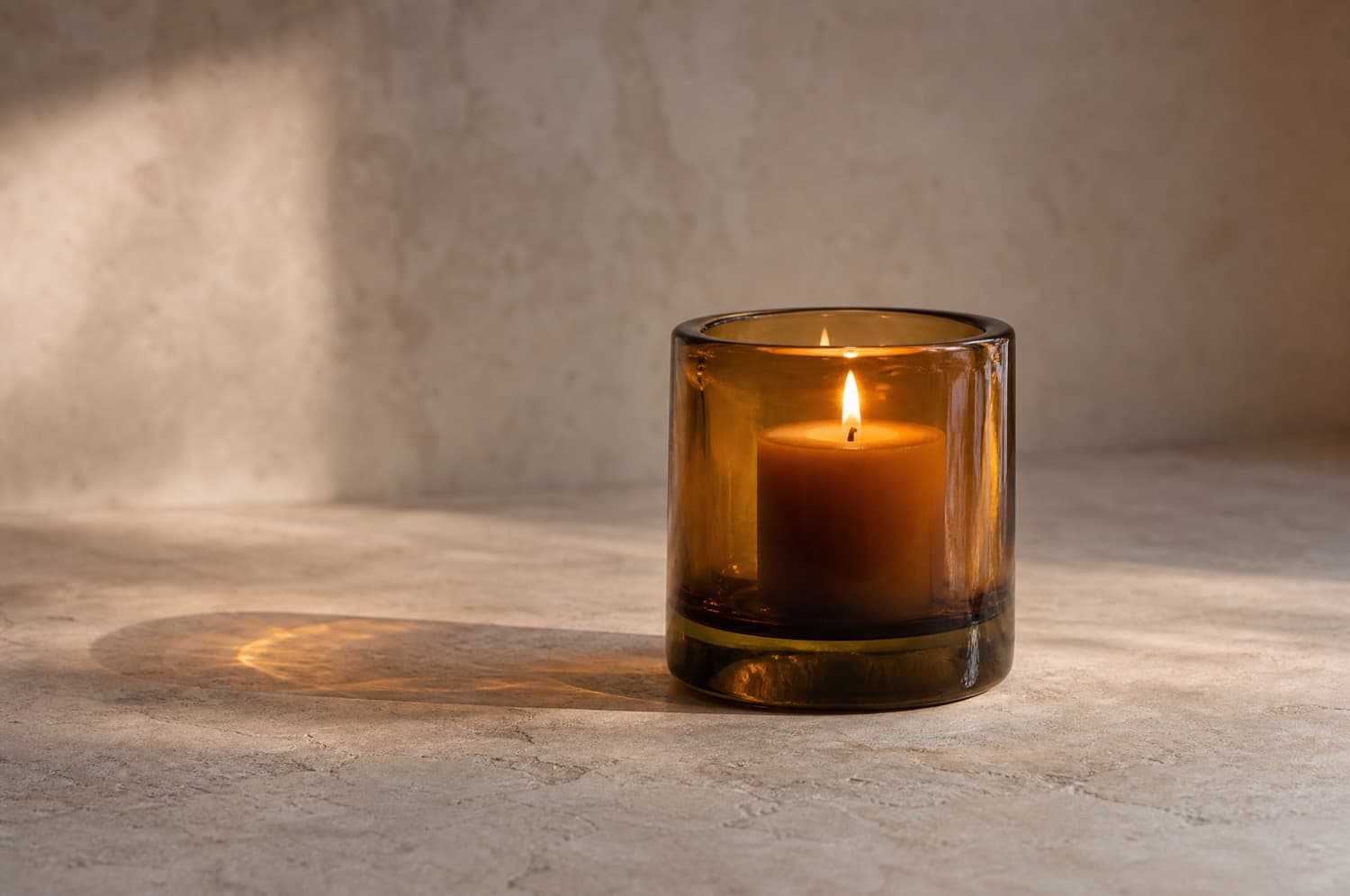A thick amber glass candle holder with a wide pillar candle burning in it on a bare stone surface, with clear space all around and nothing flammable nearby.