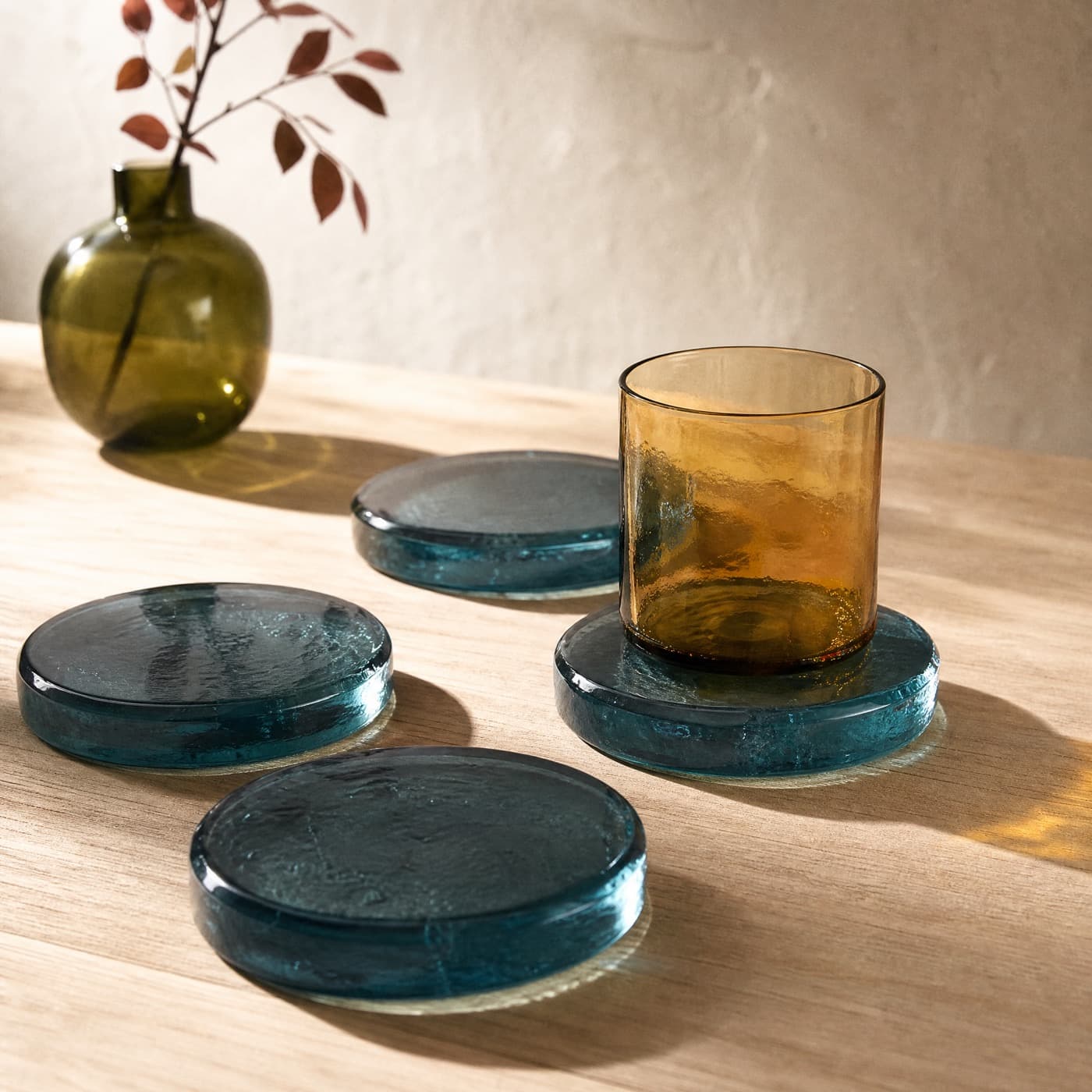 Four thick sea-blue glass coasters on an oak table, one with an amber glass standing on it.
