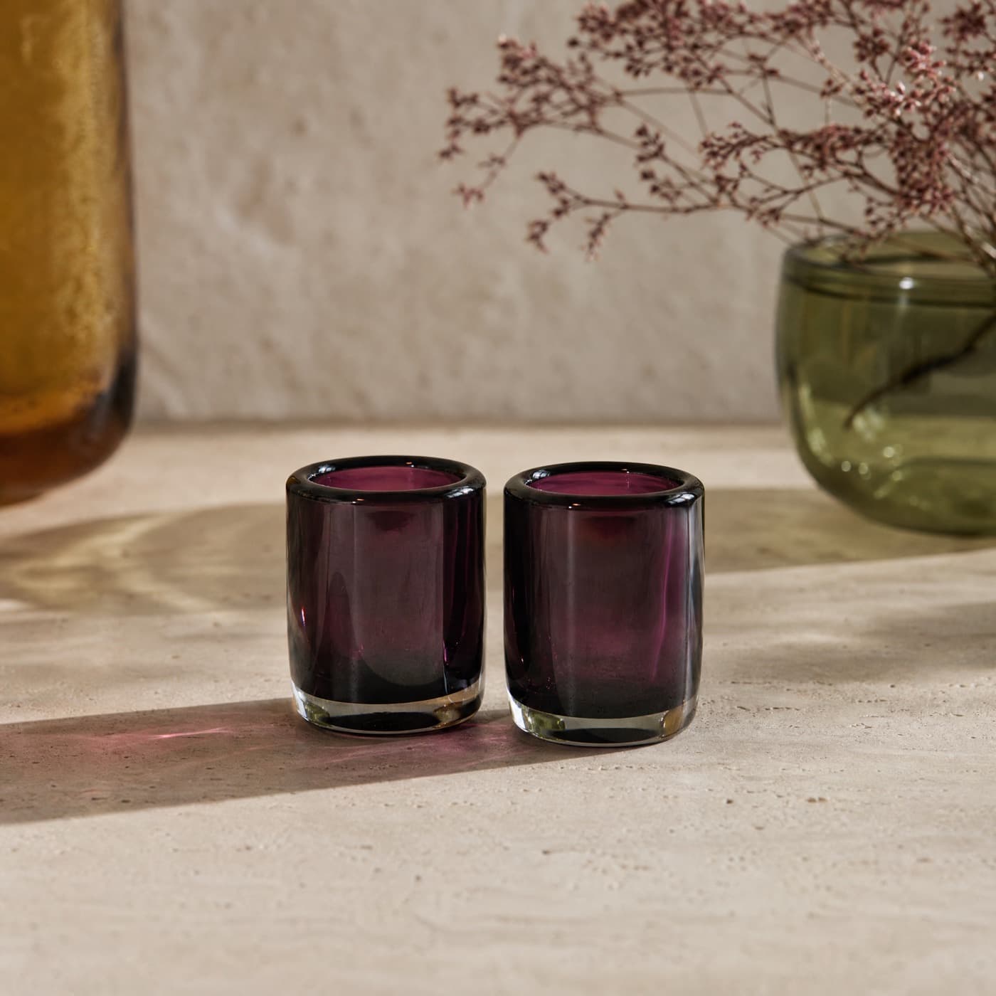 Two thick-walled plum shot glasses side by side in raking light, each dropping a dark violet patch.