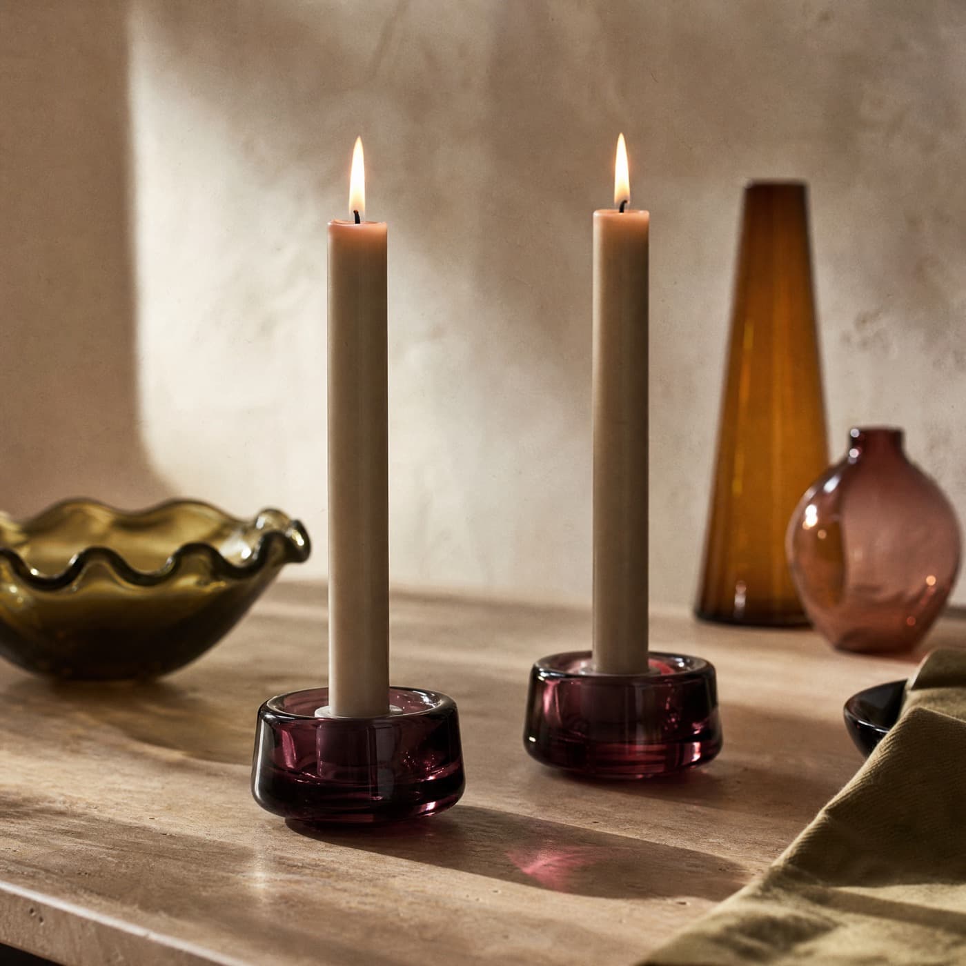 Two low plum glass taper holders with lit dinner candles on a dark evening table.